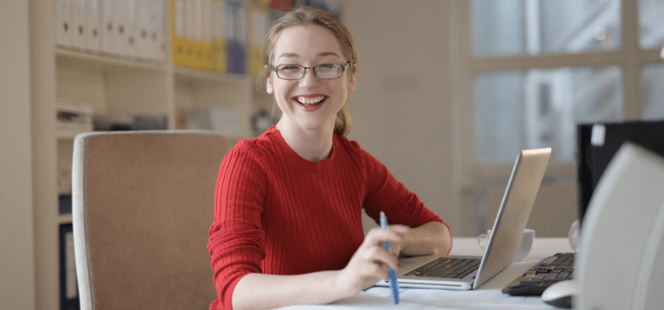 Akademia Inwestowania Alternatywnego - uśmiechnięta dziewczyna w czerwonym swetrze i okularach siedzi przy biurku z laptopem
