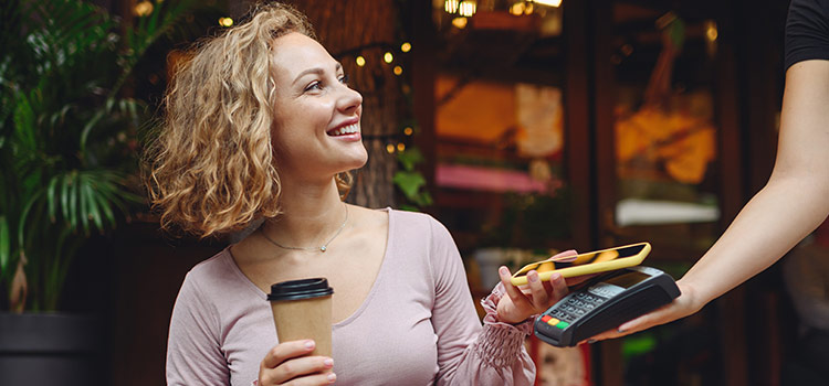 Uśmiechnięta kobieta z kubkiem kawy na wynos płaci zbliżeniowo telefonem przy terminalu